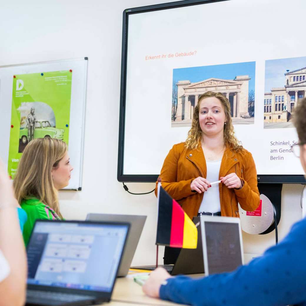 Student leraar Duits in oranje/bruin jasje geeft een lesje aan haar medestudenten. Ze staat voor een PowerPoint.
