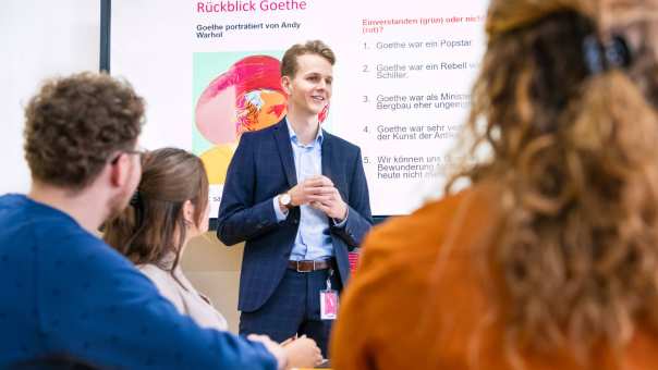 Leraar Duits in pak staat voor de klas met zijn handen in elkaar gevouwen en legt iets uit. Hij heeft een Powerpoint op de achtergrond openstaan.
