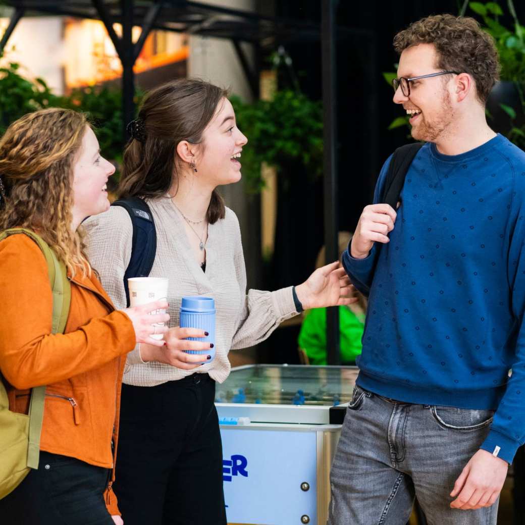 Drie studenten Leraar Duits praten met elkaar en één iemand vertelt met enthousiasme iets aan de rest. De andere twee lachen.
