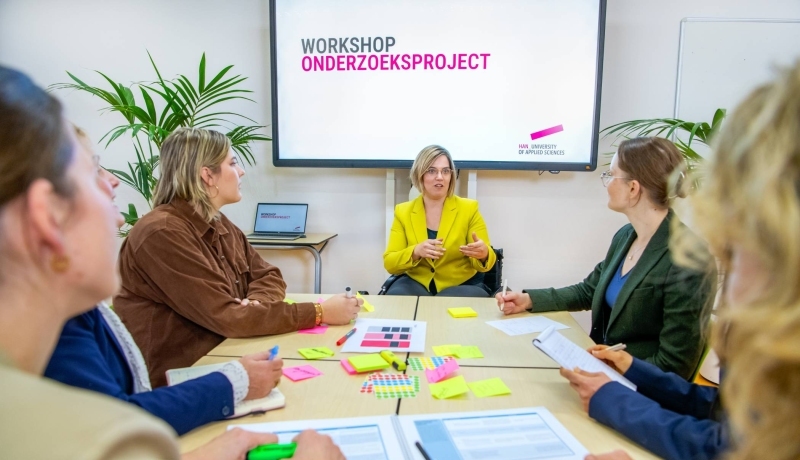 Een onderzoeker presenteert voor zes andere studenten en onderzoekers aan een tafel