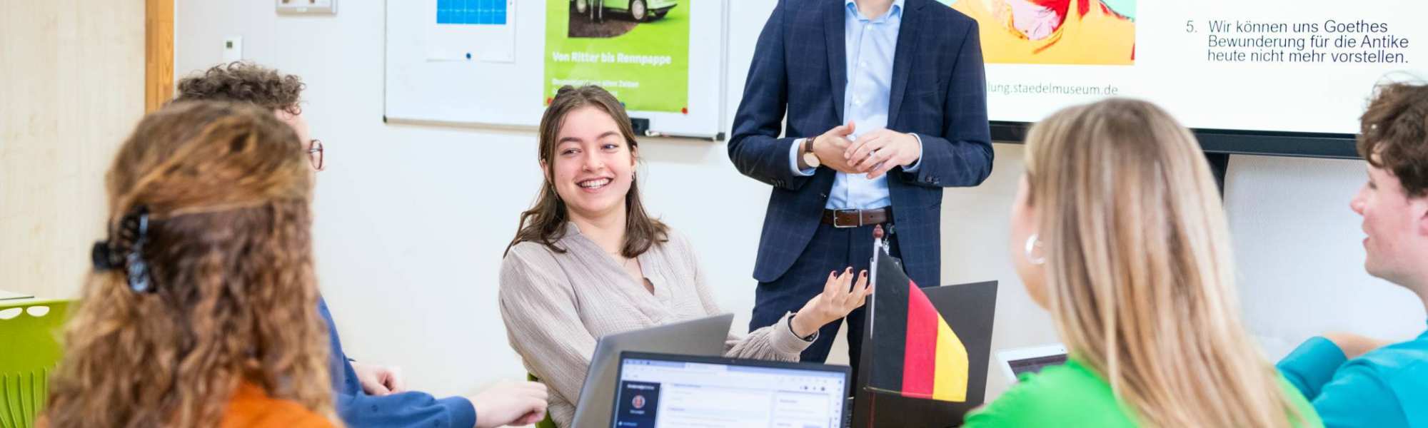 Studenten zitten samen aan tafels met laptops en boeken in het klaslokaal. De leraar staat ervoor en praat met hen mee.
