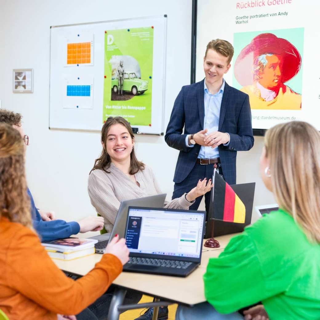 Studenten zitten samen aan tafels met laptops en boeken in het klaslokaal. De leraar staat ervoor en praat met hen mee.
